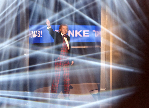 Ein Moderator winkt auf einer Bühne zwischen hellen Lichtstrahlen.